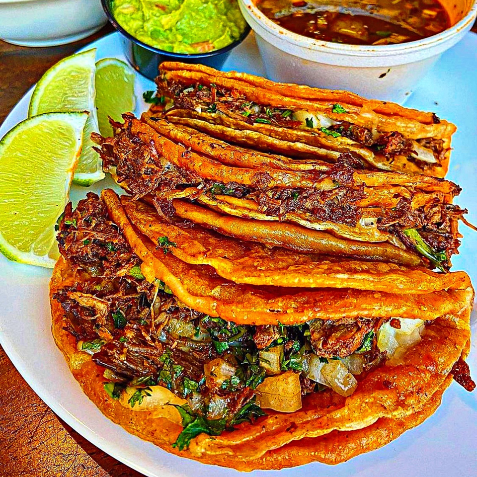 A plate of El Agave Azul Baymeadows birria tacos with lime wedges, guacamole, and consommé.