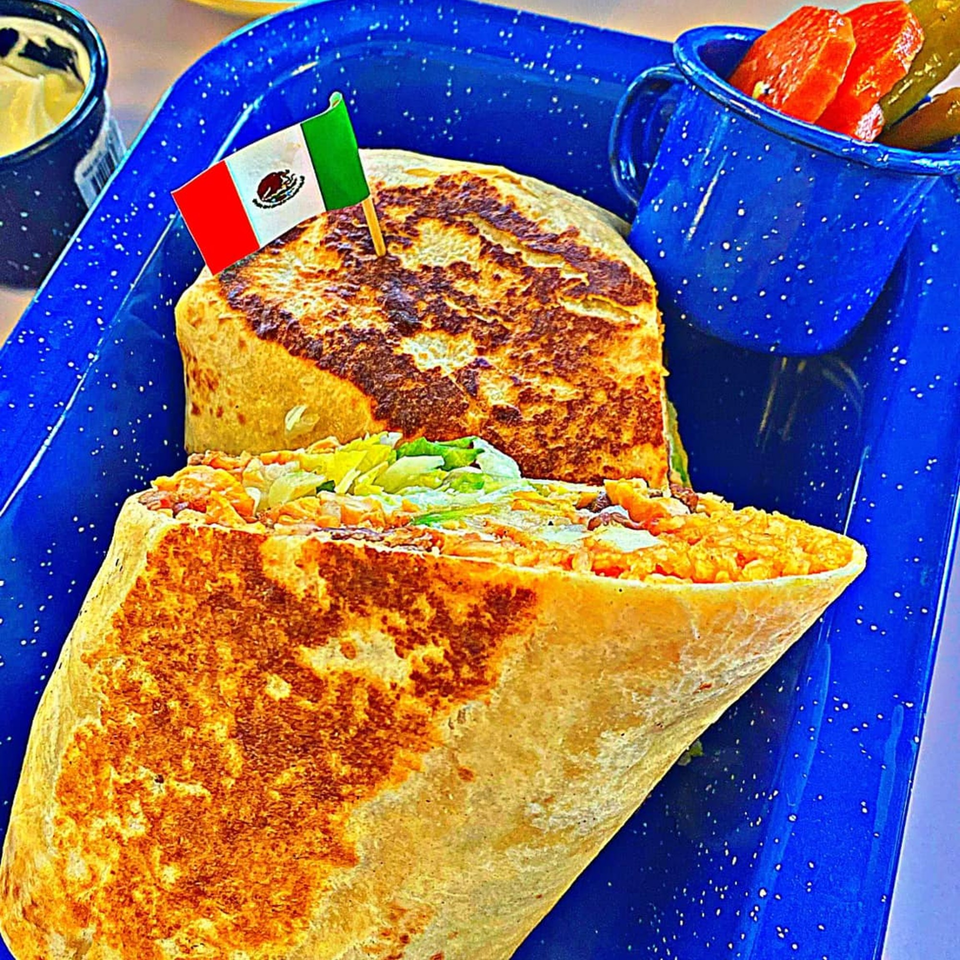 A large, grilled burrito with a Mexican flag toothpick, served with pickled vegetables on a blue tray.