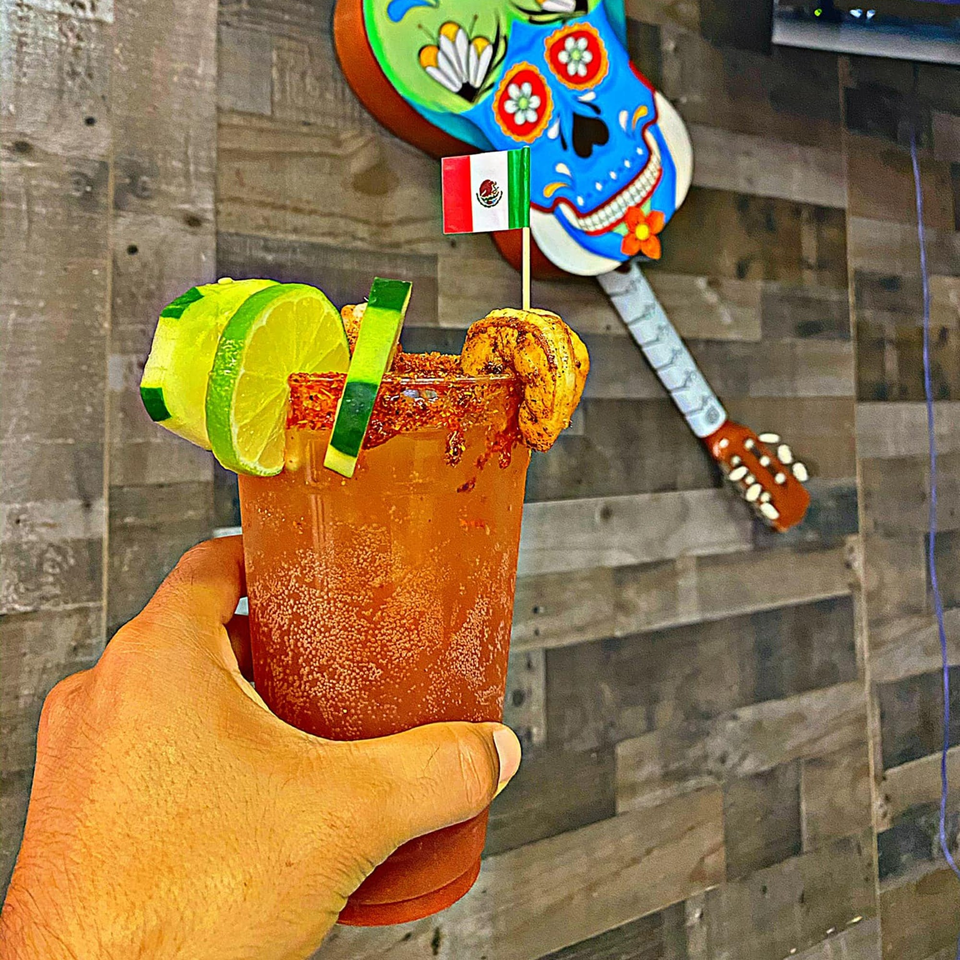 Hand holding a michelada with lime, cucumber, shrimp, and a Mexican flag, against a wooden wall.